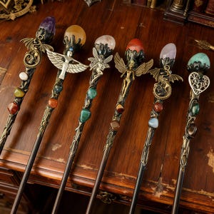 K&ouml;nnte beinhalten: Eine Sammlung von sechs verzierten Fantasy-Zauberst&auml;ben. Jeder Zauberstab hat einen dunklen Holzschaft, verziert mit dekorativen Metallarbeiten und farbigen Edelsteinen. Die Spitzen sind mit verschiedenen Kristallkugeln und detaillierten Figuren verziert, darunter Drachen, Feen und ein Engel.