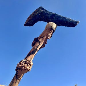 Peut inclure: Une hache en obsidienne noire fixée à un manche en bois avec une corde brune enroulée autour de la jointure. Le manche est en bois naturel avec des nœuds et une finition brun foncé.