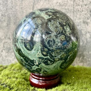 May include: A polished, spherical gemstone with a swirling pattern of dark green and black, resting on a small, round, dark red wooden stand. The sphere is displayed on a bed of green moss.