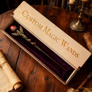 K&ouml;nnte beinhalten: Eine individuelle Zauberstab in einer offenen Holzkiste. Der Zauberstab hat einen roten Edelstein und eine goldfarbene Feenfigur. Das Innere der Box ist mit violettem Samt ausgekleidet. Auf dem Deckel steht "Custom Magic Wands".