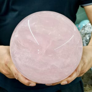 May include: A large, polished rose quartz sphere, held in both hands. The sphere is a soft pink color with subtle internal veining and a smooth, reflective surface. The background is a dark blue shirt.