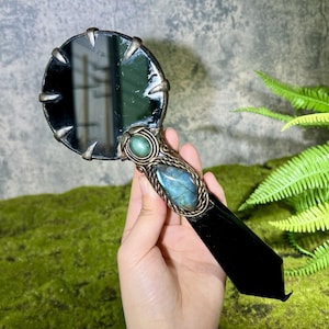 Puede incluir: Un espejo de mano con una superficie de cristal circular sujeta por garras metálicas. El mango está adornado con una piedra ovalada verde lisa y una labradorita azul iridiscente más grande, engastadas en un marco metálico oscuro y texturizado.
