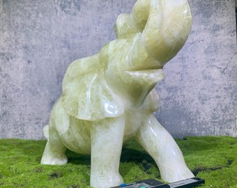 May include: A light green elephant sculpture with its trunk raised. The figurine is made of a translucent material. A digital caliper is in the foreground, possibly for measurement. The elephant is on a green surface.