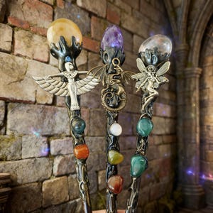 K&ouml;nnte beinhalten: Drei dekorative Fantasy-Zauberst&auml;be mit kunstvollen Designs. Jeder Zauberstab hat einen Edelstein an der Spitze und aufwendige Metallarbeiten. Ein Zauberstab hat eine Anubis-Figur, ein anderer einen Drachen und der dritte eine Fee. Die Zauberst&auml;be stehen vor einer Backsteinmauer.