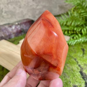 May include: A red carnelian crystal point with a smooth, polished surface. The crystal has a deep red colour with orange and white streaks throughout.