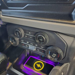 May include: Interior view of a car dashboard with a focus on the climate control and infotainment system. The controls have textured knobs. A wireless charging pad with a yellow lightning bolt symbol is visible.