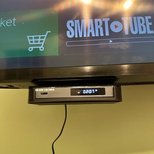 May include: A black "ELITE ULTRA" streaming device is mounted below a television screen. The device displays "82:03" on its digital display. The TV screen shows the words "SMART TUBE" and a shopping cart icon.