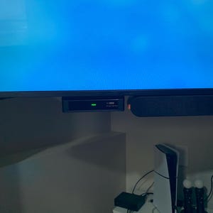 May include: A black rectangular media device with a single green indicator light mounted beneath a television screen. Below, a white gaming console stands vertically next to a black charging dock with two controllers and a small black box.