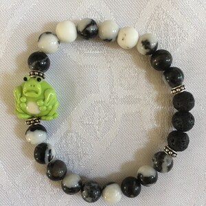 May include: A black and white stone bracelet with a green frog charm. The bracelet is made with lava rock and black and white zebra jasper beads.