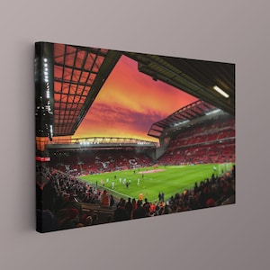 Könnte beinhalten: Leinwanddruck eines Fußballstadions bei Sonnenuntergang. Der Himmel ist eine lebendige Mischung aus Orange- und Rottönen. Das Stadion ist mit Zuschauern gefüllt, und das Spielfeld ist grün. Die Architektur des Stadions ist sichtbar, mit seinem Dach und den Sitzbereichen.
