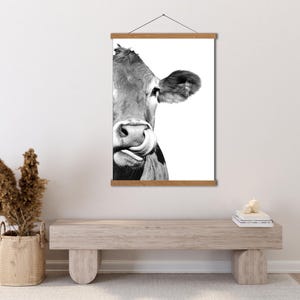 May include: Monochrome print of a cow licking its nose, mounted on a wooden frame with a hanging cord. The cow's face is central, set against a white backdrop. A rustic, farm-themed artwork.