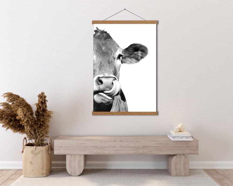 May include: Monochrome print of a cow licking its nose, mounted on a wooden frame with a hanging cord. The cow's face is central, set against a white backdrop. A rustic, farm-themed artwork.