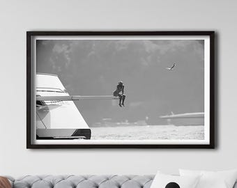 May include: A black and white framed wall art print featuring a person sitting on the diving board of a yacht. The yacht has the name JONIKAL visible on its side. A bird is flying in the sky above the water in the background.