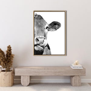 May include: Monochrome framed print featuring a cow's face, with its tongue extended. The cow's ear and eye are visible. The print is in a gold frame and is displayed on a white wall.