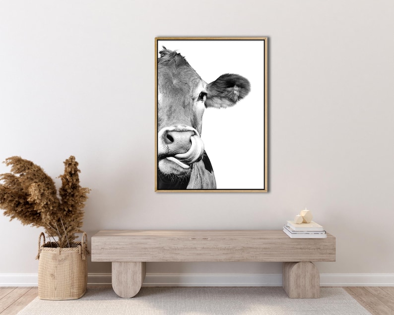 May include: Monochrome framed print featuring a cow's face, with its tongue extended. The cow's ear and eye are visible. The print is in a gold frame and is displayed on a white wall.
