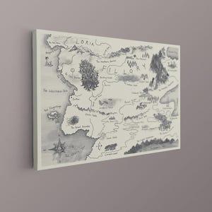 May include: A rectangular wall art print featuring a black and white hand-drawn map of the fictional land of Fillory and Loria. The map includes detailed illustrations of mountains, forests, and bodies of water, with various locations labelled in black ink.