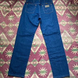 May include: Dark blue denim jeans with the Wrangler logo on the back pocket. The jeans have a classic five-pocket design with brass rivets and a straight leg. The jeans are displayed on a patterned fabric.