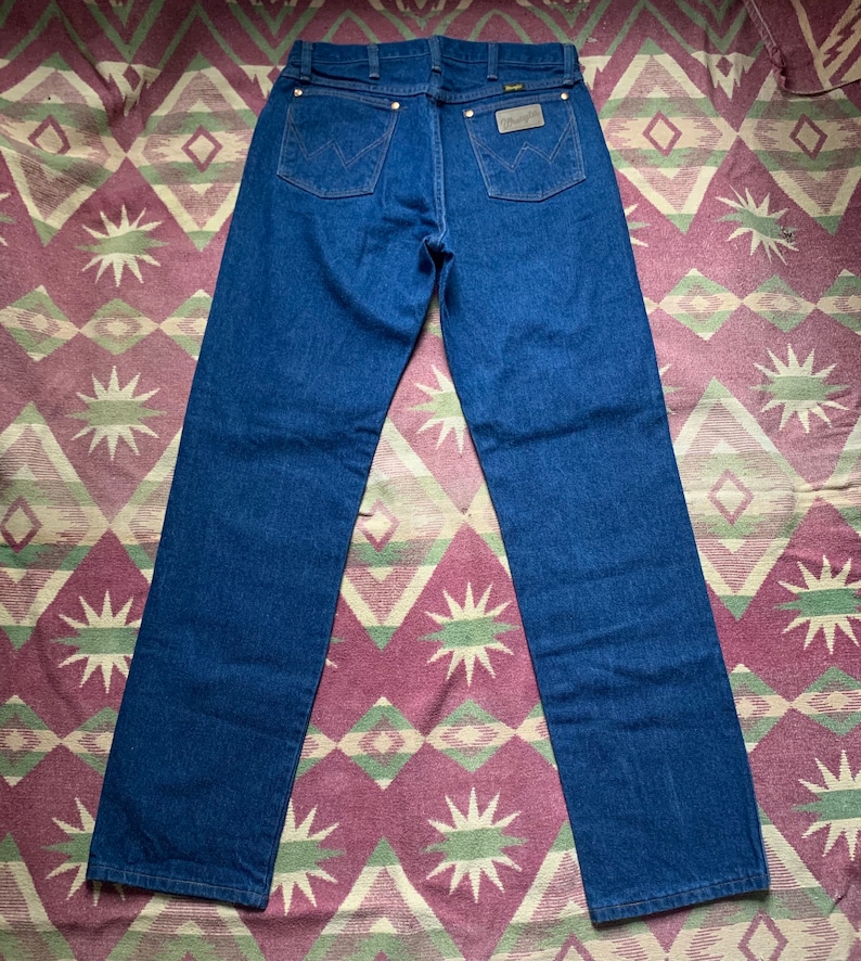 May include: Dark blue denim jeans with the Wrangler logo on the back pocket. The jeans have a classic five-pocket design with brass rivets and a straight leg. The jeans are displayed on a patterned fabric.