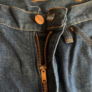 May include: Close-up of the front of a pair of dark blue denim jeans. The zipper is open, revealing the brass-colored metal zipper teeth. A copper-colored button with the brand name "Wrangler" is visible.
