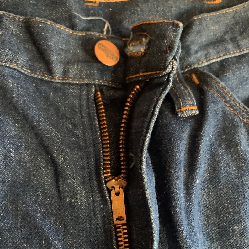 May include: Close-up of the front of a pair of dark blue denim jeans. The zipper is open, revealing the brass-colored metal zipper teeth. A copper-colored button with the brand name "Wrangler" is visible.