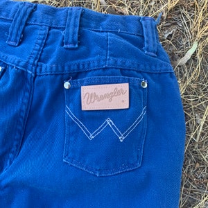 May include: Close-up of the back of blue Wrangler jeans. The jeans feature a light brown leather patch with the Wrangler logo above the back pocket. White stitching outlines the pocket and the logo patch. Silver rivets are visible.