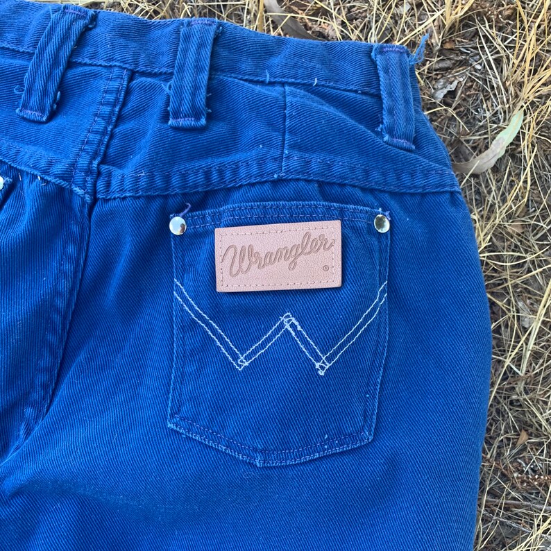 May include: Close-up of the back of blue Wrangler jeans. The jeans feature a light brown leather patch with the Wrangler logo above the back pocket. White stitching outlines the pocket and the logo patch. Silver rivets are visible.