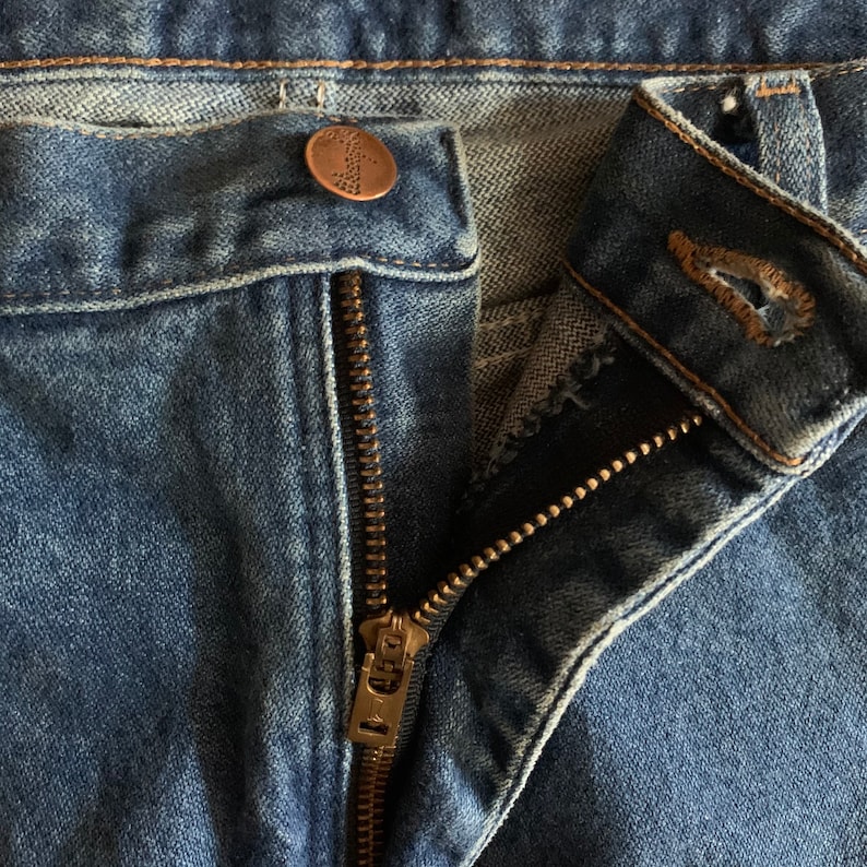 May include: Close-up of blue denim jeans with a partially unzipped brass zipper. The jeans have a button closure at the waist and a button with a decorative design. The fabric shows signs of wear.