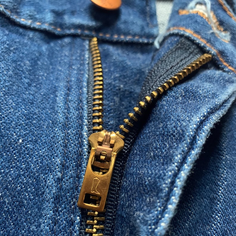 May include: Close-up of a blue denim jeans zipper. The zipper is gold-colored and partially open, revealing the teeth and the fabric of the jeans. The jeans have a copper-colored button at the top.