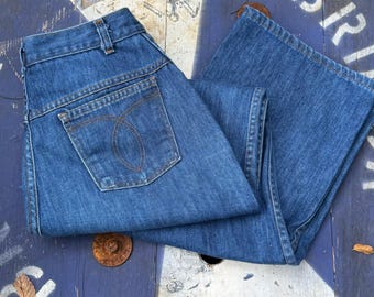 May include: Blue denim jeans with a classic five-pocket design. The jeans feature a high waist and a straight leg cut. The back pocket has a decorative stitch design. The jeans are folded and displayed on a weathered blue and white surface.