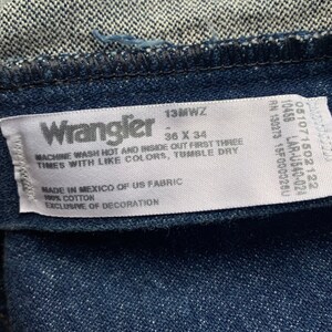 May include: Close-up of a Wrangler jeans label. The label is white with black text, including the brand name, size 36 x 34, and care instructions. The jeans are a dark blue denim.