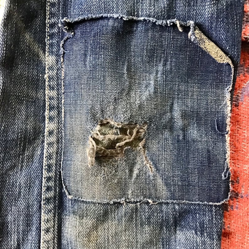 May include: Close-up of a distressed denim patch sewn onto blue jeans. The patch has a frayed edge and a large hole, revealing the texture of the fabric underneath. The jeans show signs of wear and tear.