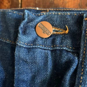May include: Close-up of a pair of dark blue denim jeans. The jeans feature a copper-colored button with the brand name "Wrangler" embossed on it. The button is secured to the jeans with a matching copper-colored thread.
