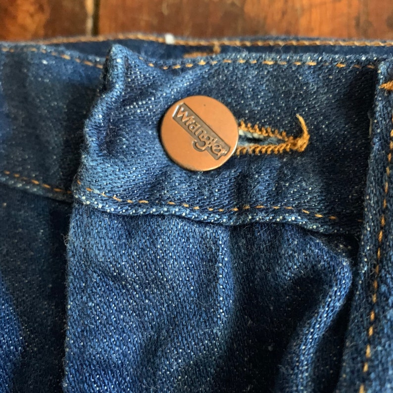 May include: Close-up of a pair of dark blue denim jeans. The jeans feature a copper-colored button with the brand name "Wrangler" embossed on it. The button is secured to the jeans with a matching copper-colored thread.