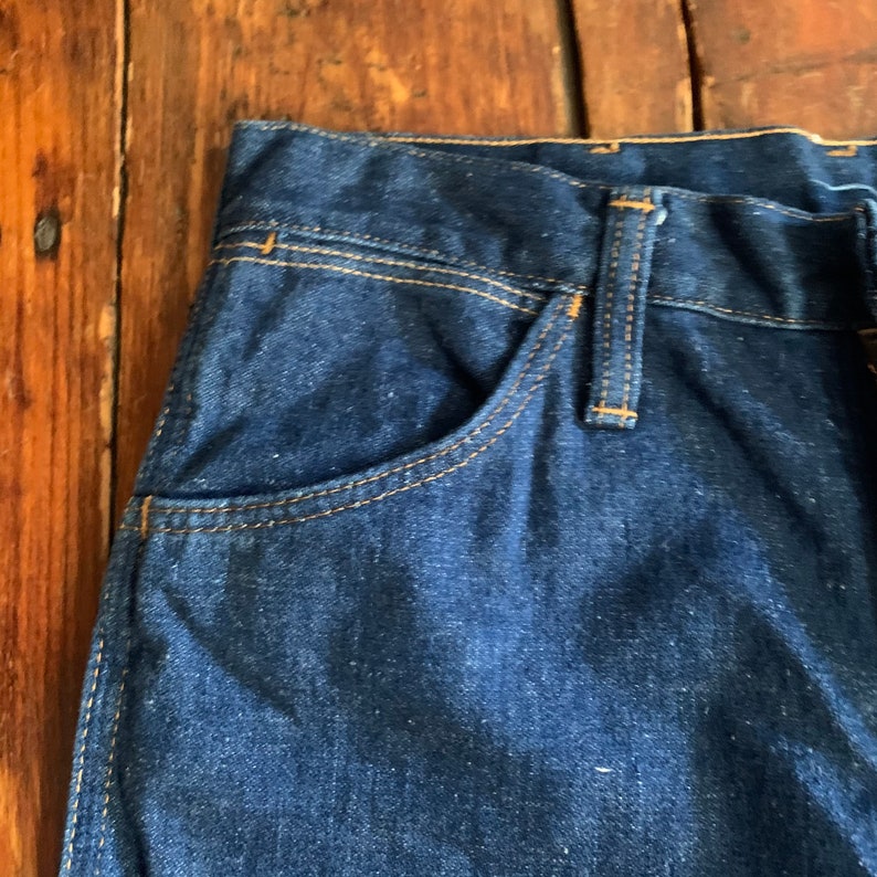 May include: Close-up of dark blue denim jeans. The jeans feature a classic five-pocket design with a front pocket and a belt loop. The stitching is a contrasting tan color.