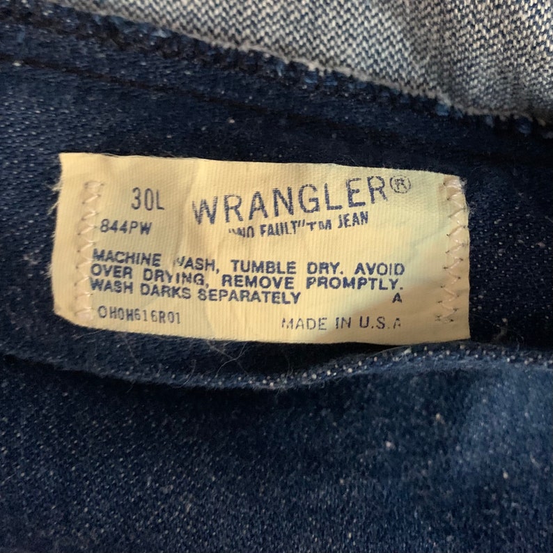 May include: Close-up of a Wrangler jeans tag. The tag is off-white with black text, including the brand name, size 30L, and care instructions. The tag reads "NO FAULT"TM JEAN" and "MADE IN U.S.A."