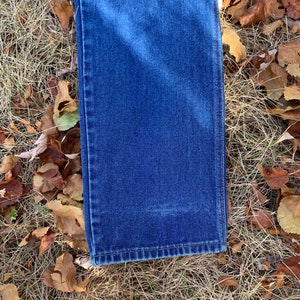 Pu&ograve; includere: Primo piano di un paio di jeans blu in denim, piegati per mostrare l'orlo e la trama del tessuto. I jeans sono appoggiati su erba secca e foglie cadute. Il denim &egrave; di un blu intenso.