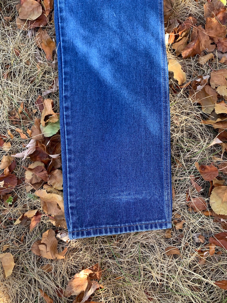 Pu&ograve; includere: Primo piano di un paio di jeans blu in denim, piegati per mostrare l'orlo e la trama del tessuto. I jeans sono appoggiati su erba secca e foglie cadute. Il denim &egrave; di un blu intenso.