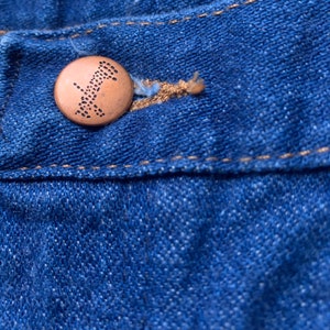 May include: Close-up of blue denim fabric with a copper-colored button. The button has a detailed design and is attached to the fabric with a tan-colored thread. The denim has visible stitching.