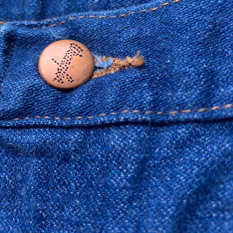 May include: Close-up of blue denim fabric with a copper-colored button. The button has a detailed design and is attached to the fabric with a tan-colored thread. The denim has visible stitching.