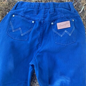 May include: Close-up of the back of a pair of blue Wrangler jeans. The jeans have two back pockets with white stitching in a W pattern. The Wrangler logo is on a brown leather patch. Silver rivets are visible.