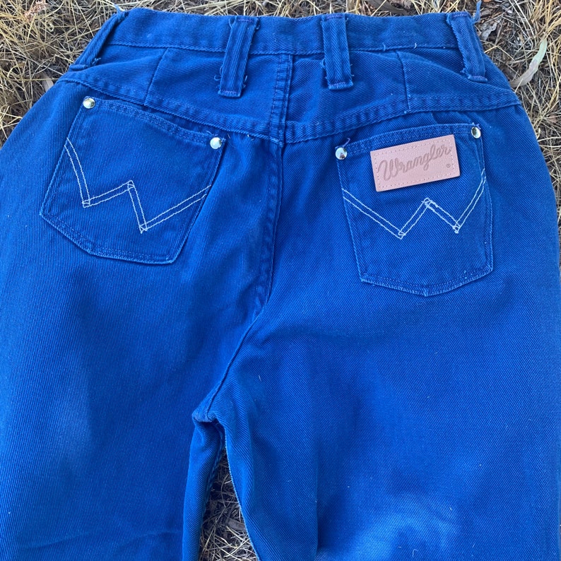 May include: Close-up of the back of a pair of blue Wrangler jeans. The jeans have two back pockets with white stitching in a W pattern. The Wrangler logo is on a brown leather patch. Silver rivets are visible.