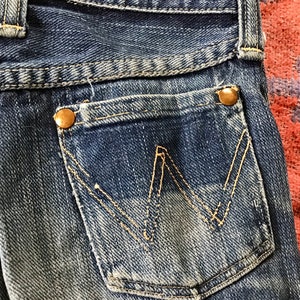 May include: Close-up of a pair of blue denim jeans. The back pocket features a distinctive 'W' shaped stitching in a contrasting yellow thread. The pocket is secured with a brass-colored button. The jeans show signs of wear.
