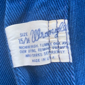 May include: Close-up of a blue fabric with a white label. The label reads "Wrangler" in cursive, with size 15/16. Washing instructions are also printed on the label. Made in the USA.