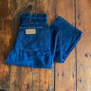 May include: Dark blue denim Wrangler jeans with a brown leather patch on the back pocket. The jeans are folded and displayed on a wooden surface. The Wrangler logo is visible on the patch.