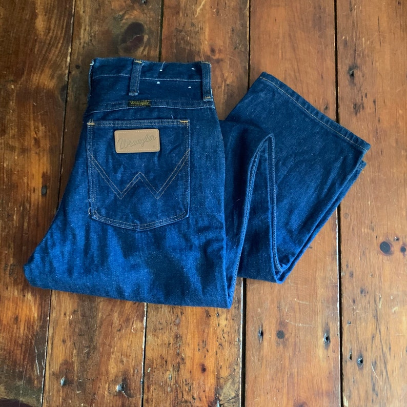 May include: Dark blue denim Wrangler jeans with a brown leather patch on the back pocket. The jeans are folded and displayed on a wooden surface. The Wrangler logo is visible on the patch.
