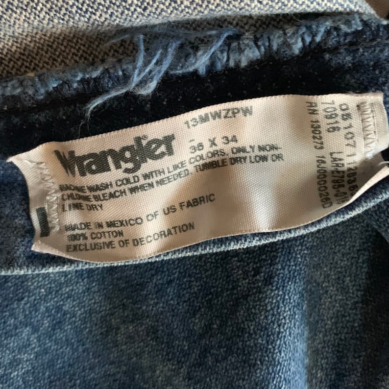 May include: Close-up of a Wrangler jeans tag. The tag is white with black text, showing the brand name, size 36 x 34, and care instructions. The jeans are a medium-wash blue denim.