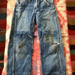 May include: Blue denim jeans with patched knees. The jeans have a classic five-pocket design, a button and zipper closure, and belt loops. The denim has a faded, worn appearance.