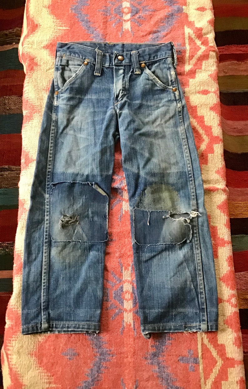 May include: Blue denim jeans with patched knees. The jeans have a classic five-pocket design, a button and zipper closure, and belt loops. The denim has a faded, worn appearance.