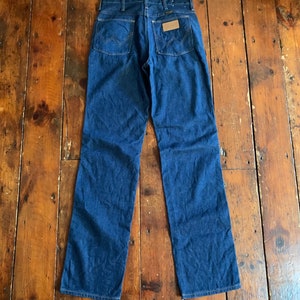 May include: Dark blue denim jeans with a classic five-pocket design. The jeans feature the iconic Wrangler stitching on the back pockets and a brown leather patch. The jeans are laid flat on a wooden surface.