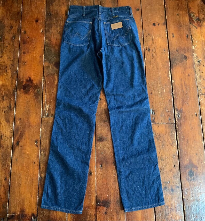 May include: Dark blue denim jeans with a classic five-pocket design. The jeans feature the iconic Wrangler stitching on the back pockets and a brown leather patch. The jeans are laid flat on a wooden surface.